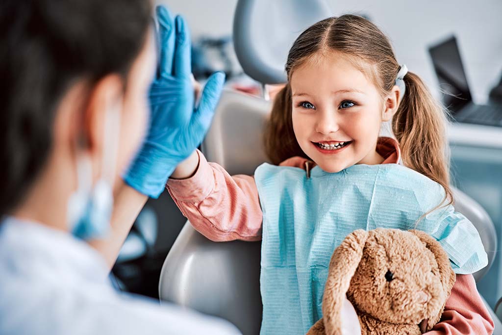 Young girl holding a stuffed rabbit a the pediatric dentist high fives her practitioner and smiles, demonstrating the value of going to a pediatric dentist