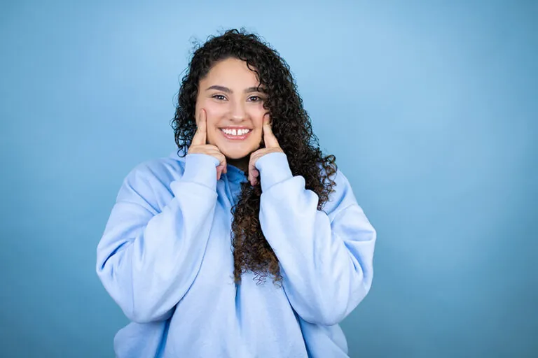 Young woman with dark curly hair and a gap in her front teeth wears a light blue sweater set against a blue background